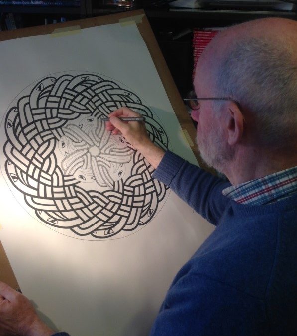 Dr Larry Scrimgeour sat at a desk working on a celtic knot. Larry is wearing a blue jumper and glasses.